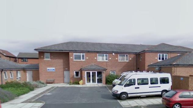 The Aire Court building in Middleton, Leeds, is a two-storey red brick construction with a car park outside the entrance. Three minibus vehicles are parked outside along with a small red car. 