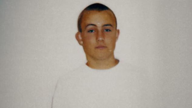 Police photograph of a teenage boy looking blanky at the camera against a white wall. He has short blond cropped hair and is wearing a long-sleeved white top.
