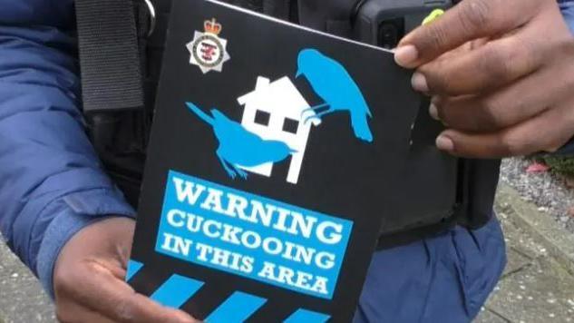 A close up of a police officer wearing a protection vest and long sleeved blue raincoat as he holds a poster saying "Warning cuckooing in this area". The poster has the Avon and Somerset Police branding on it, and a house with two blue cuckoo outlines on it.