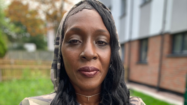 Ms Mellis is wearing a green camouflage top with the hood up and is smiling at the camera. She has a choker-type gold necklace on and has long dark hair. She is stood on grass outside a building.