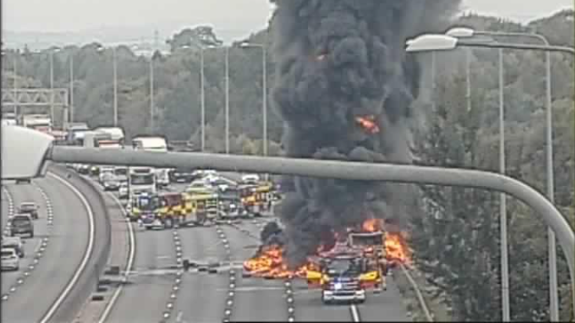 A lorry is on fire on the M25, and a fire truck is parked in front of it. There are a lot of flames and black smoke is billowing into the sky. There is traffic snarled up in the background.