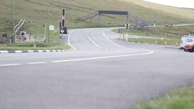 The A18 Mountain Road snaking under a pedestrian bridge over the carriageway in the direction of Douglas. The barriers and lights for the Snaefell Mountain Railway can also be seen.