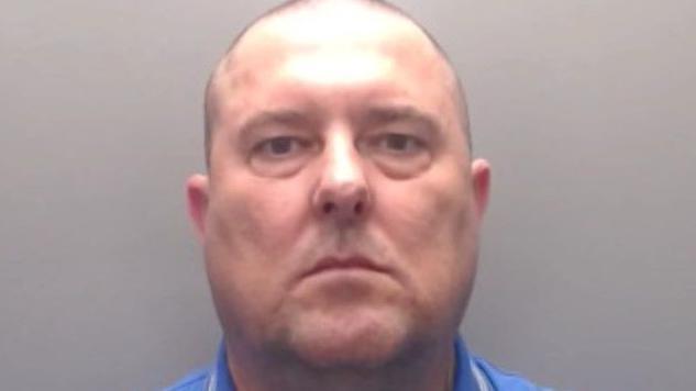 A mugshot of a bald Jason Howe who is in his 50s with tattoos on his arms wearing a blue Boss branded T-shirt.