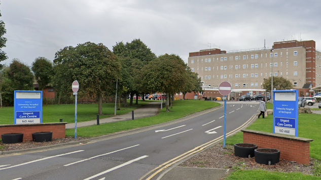 University Hospital of Hartlepool entrance with a road leading up to a large grey building. There are two blue signs on both sides of the road reading 'Urgent Care Centre' and 'No A&E'.