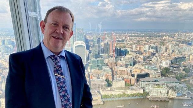 Ian Vowles Smith wears a blue suit and stands in front of a window with a cityscape behind looks at the camera