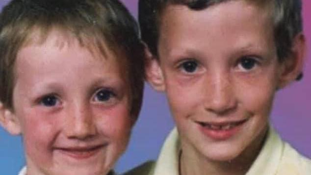 Two young boys with mousy hair and blue eyes smiling for the camera. The boy on the right is wearing a yellow top.