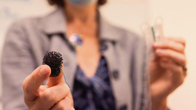 Prof Rebecca Fitzgerald in the background, wearing a mauve jacket over a purple top and with a surgical mask on. She is holding in the foreground a black sponge in her right hand which has a string attached