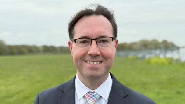 Mark Flynn wears glasses and stands in a field. He wears a navy suit jacket, light-coloured check-patterned tie and a pale blue shirt. He is smiling at the camera.