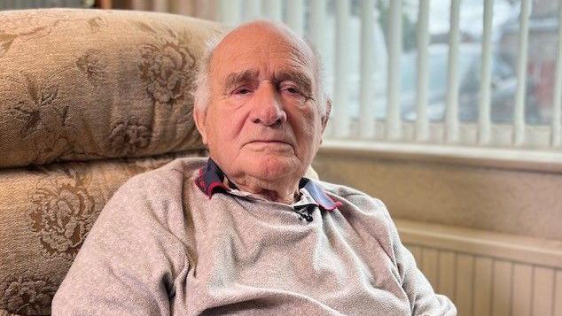 90-year-old Ted McKay in a grey jumper, at home, looking at the camera 