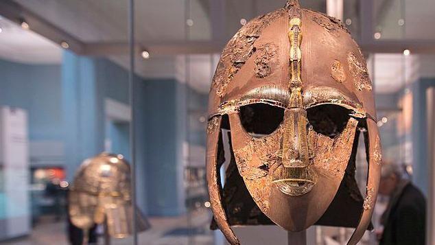 The Sutton Hoo Anglo-Saxon helmet is pictured in a glass display cabinet at the British Museum. It is a bronze coloured helmet with holes for the wearer's eyes. It has panels that would run down the side of the wearer's face and down the back of their neck. A young woman with brown hair looks at the helmet from the side.