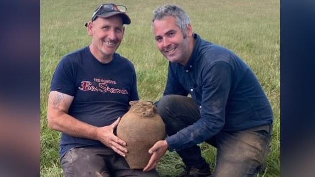 Two men holding a clay pot smiling with the dig site seen just in front of them. The man on the left has glasses on his head and a cap, he has a blue t-shirt with the words Ben Sherman, in red font, written on the front. The man on the right has salt and pepper hair, a blue shirt and blue pants.