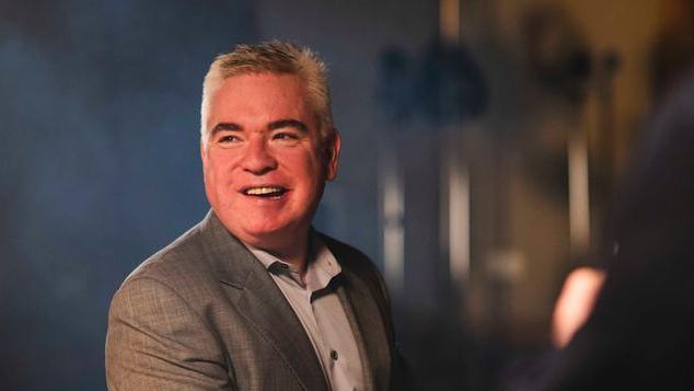 Lee Michael Walton, a man with short grey hair wearing a grey suit jacket and grey shirt, sits at a black grand piano looking off to the side of the camera and smiling