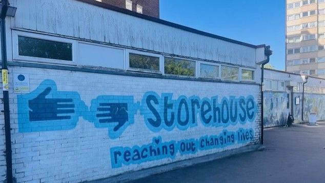 A predominately white building, with "Storehouse" and reaching out changing lives" painted in blue on the front.  Two hands are also painted in blue on the front.  There is a high rise building ion the background.  The sky is blue.