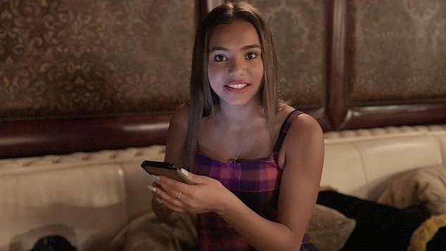 Lily sits on a white sofa and smiles at the camera while holding a phone. She is wearing tartan pink dress and has brown hair.