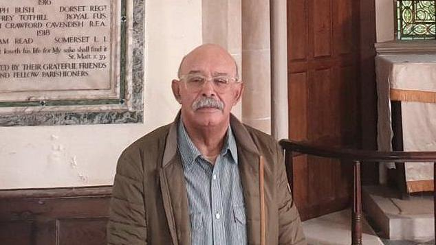 John Stockley wearing a grey shirt and brown anorak, standing in front of a war memorial at the front of a church, with the altar in the background.