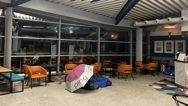 A person is sleeping on the floor of an airport cafe, the have a sleeping bag and an opened umbrella giving them some privacy 