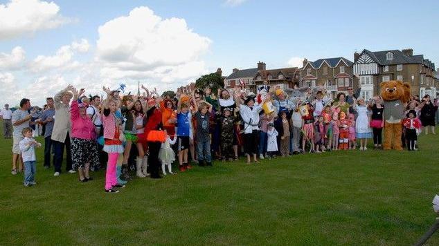 A large group of people wearing different fancy dress outfits stand at an angle waving to the photographer on a grassy common. The sky is blue with some cloud.