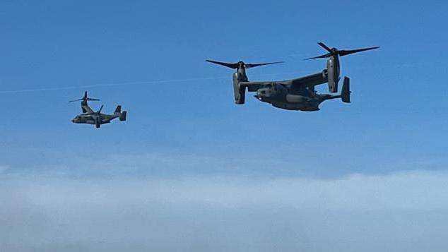 Two Osprey helicopters in a clear blue sky, with a band of white cloud beneath and a slight rainbow caused by the downdraught.