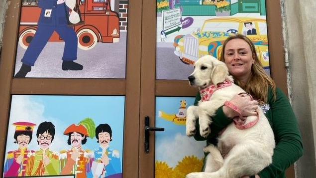 Kath McGuire, founder of the Penny Lane Wombles community group which organises regular litter picks in the area, poses for a photograph while holding her golden retriever Rita in her arms. They are photographed in front of artwork depicting The Beatles in Sgt Pepper-era suits.