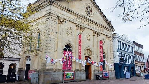 Corn Exchange Newbury is a tall, light stone building with carvings and topped by a triangular pediment. The ornate front entrance is flanked by a large arched window on either side.