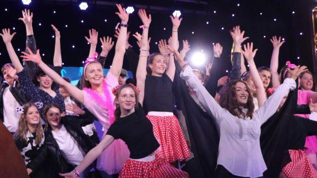 Llun yn dangos grwp o bobl mewn gwisgoedd amrywiol ar lwyfan yn dal eu breichiau i fyny, fel petai nhw newydd orffen perfformiad.