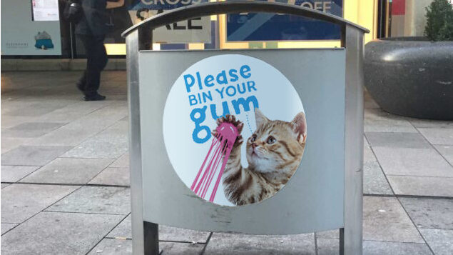 A bin with an image of a cat with chewing gum on its paw. It says 'please bin your gum' on it.