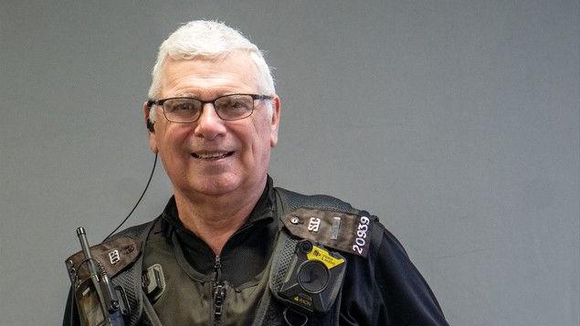 Police officer Kevin Lee is stood against a grey background. He is wearing glasses and black police uniform with a body-worn camera, police radio and other police equipment attached.