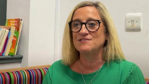 Sarah Coyle smiles while sitting on a multi coloured striped sofa, wearing a green top and large dark framed glasses. On a shelf next to her are a number of books and a little toy house 