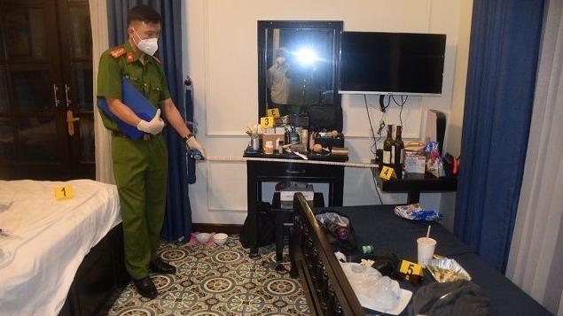 A police officer uses a tape measure while standing by a bed in a villa. Make up sits on a dresser, and food and drink containers sit on a side table and sofa. Yellow crime scene placards are dotted around.