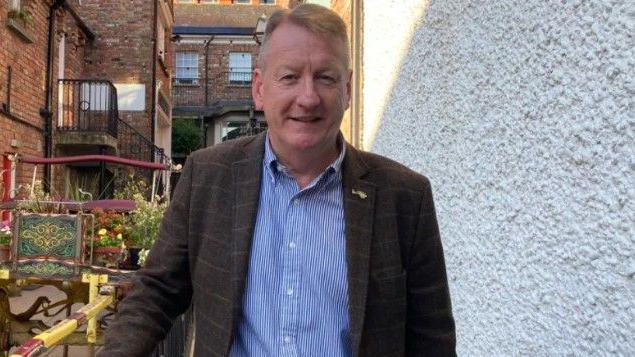 Jim Roddy wearing a brown checked jacket and a blue striped button-down shirt with a whitewashed wall to his left