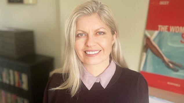 A woman with blonde hair staring at the camera and smiling. She is wearing a dark jumper with a pink shirt collar around the neck