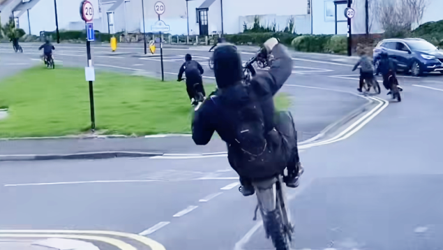 Six people on bikes on a Tredegar road, with one in the forefront with hood up and doing a wheelie, with the front wheel high in the air