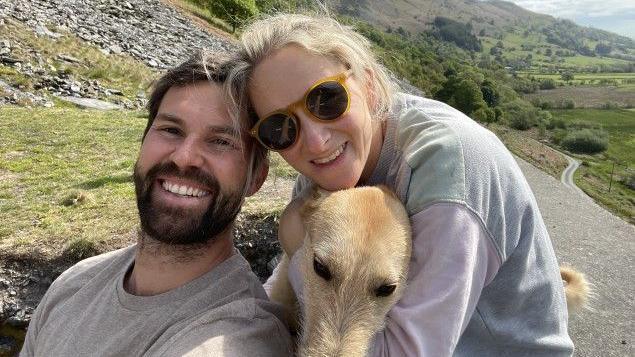 A man and a woman smile at the camera, as a woman hugs a golden dog. The trio are in front of rolling green hills and a road.