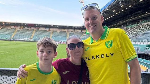 Jason with his wife and son, wearing Canaries football shirts. They are all standing inside of a football stadium.