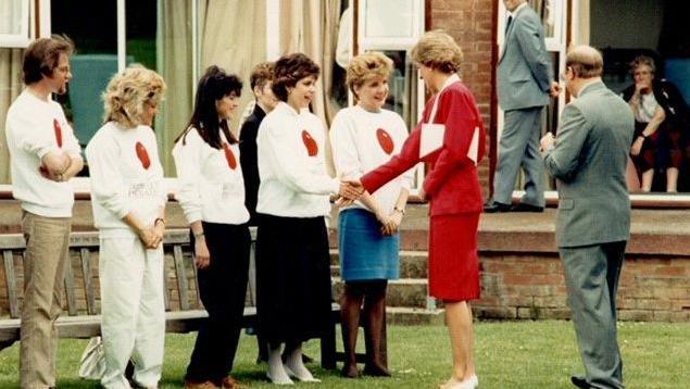 Katie shaking hands with Princess Diana at Papworth Hospital. Katie is a line with other people. Princess Diana is wearing a red skirt and matching jacket. 
