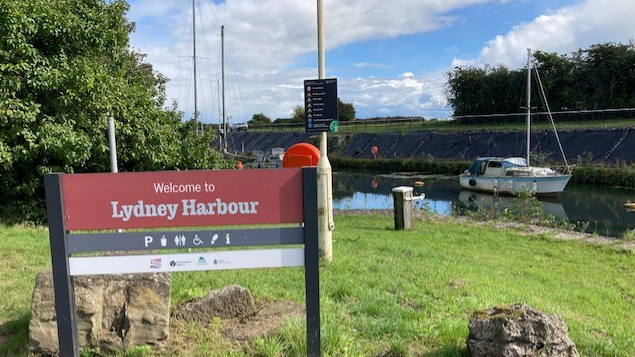 A sign on grass says 'welcome to Lydney Harbour' and there is a boat on water to the far right of the image. 