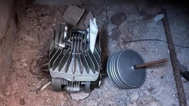 This picture shows a small cavity beneath a floor under floorboards, where a motorbike engine has been stored. The engine appears to be a single-cylinder unit with cooling fins. Next to it is a round metal object with a rod attached. There is also a folded piece of paper inserted into the engine fins. The surrounding area is rough concrete and brickwork.