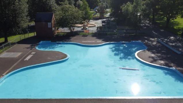 An aerial shot of a large, curved, paddling pool in a park, surrounded by grey non-slip surfaces. A children's play park and slide cam be seen in the background.