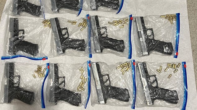 11 small black guns all contained inside zip lock bags with gold bullets. They are laid out on a white sheet on the floor.