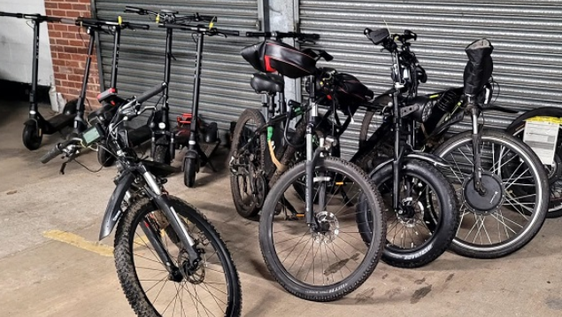 A number of black e-scooters and e-bikes in front of a metal shuttered entrance