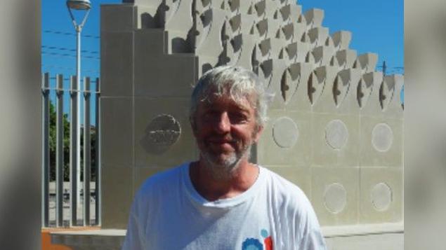 Rob Macdonald is standing in front of the Solidarity Park living monument in Spain. He is wearing a white t-shirt with a Solidarity Park logo on it and has white hair and a beard. The sculpture is made of white stones and resembles a hip. 