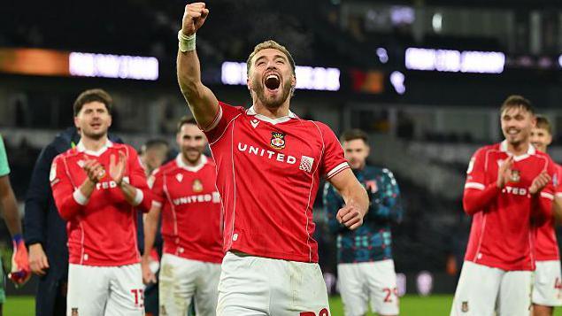 Wrexham striker Sam Smith punches the air in celebration with teammates applauding in the background