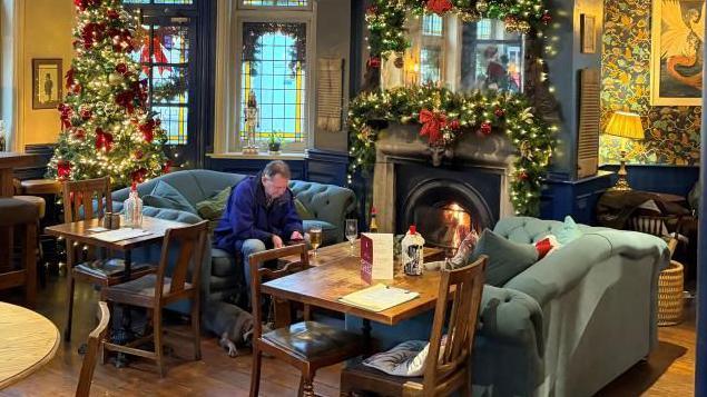 The pub is furnished with a mixture of pale green sofas and wooden tables and chairs. The fireplace is covered in a illuminated garland and a Christmas tree sits in the background. A man sits on a sofa with a stemmed glass of beer in front of him and a dog lying at his feet in front of an open fire.