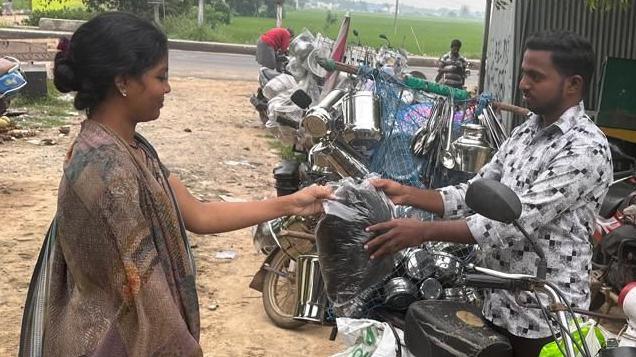 A lady hands a bag of hair to Kolachi Venkatesh who is standing next to his scooter.