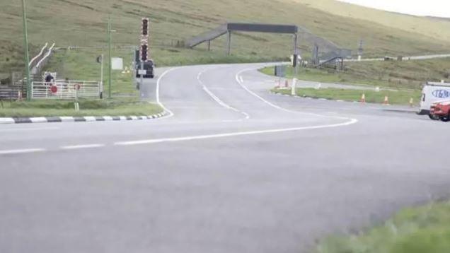 The A18 Mountain Road snaking off into the distance under a metal footbridge. Signals and gates for the Snaefell Mountain Railway can been seen on the left hand side of the road.