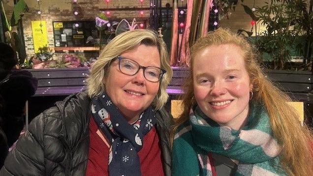 Two women smile. Both are wearing scarves. One has blonde hair and one has ginger hair. 