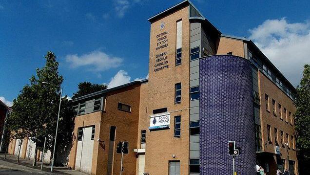 Swansea Central Police Station main entrance seen from the street. The building is light brown brick with a blue rounded facade on the corner and a large police/heddlu sign