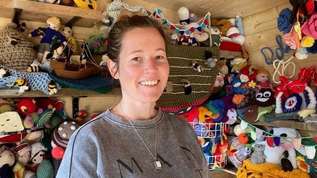 Alison Davis smiles as she stands inside a wooden shed where shelves are piled high with colourful knitted "toppers". She has dark hair, which is tied back, and wears a grey sweater and a silver necklace with white pendant.