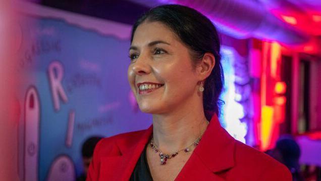 Llun yn dangos menyw efo gwallt yn tywyll a siaced coch yn edrych i ffwrdd o'r camera.