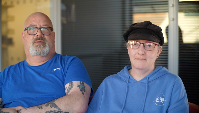 Shona sits on the right with a blue hoody and hat on, wearing red-rimmed glasses. Next to her is a man named Chris who wears a blue T-shirt, has tattoos on his arms and also has glasses on